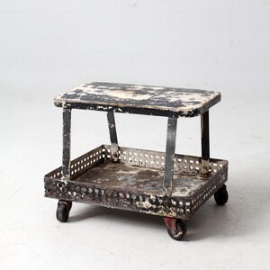 May include: A vintage industrial cart with a distressed black and white painted top and a perforated metal tray. The cart has a black frame and rolls on small wheels.
