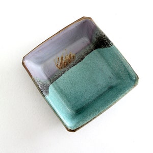 Può includere: Un piatto quadrato in ceramica con una glassa viola chiaro sulla metà superiore e una glassa turchese sulla metà inferiore. Il piatto ha un bordo marrone e un disegno marrone al centro.