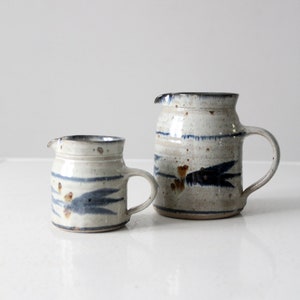 May include: Two ceramic pitchers with a white glaze and blue and brown painted designs. The smaller pitcher is on the left and the larger pitcher is on the right.