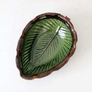 May include: A ceramic leaf-shaped dish with a detailed green leaf design in the center. The dish has a dark brown, textured rim. The overall shape is oval, and the dish is set against a white background.