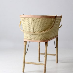 May include: A vintage wicker baby bassinet with a light pink trim and wooden legs. The bassinet is on a white background.