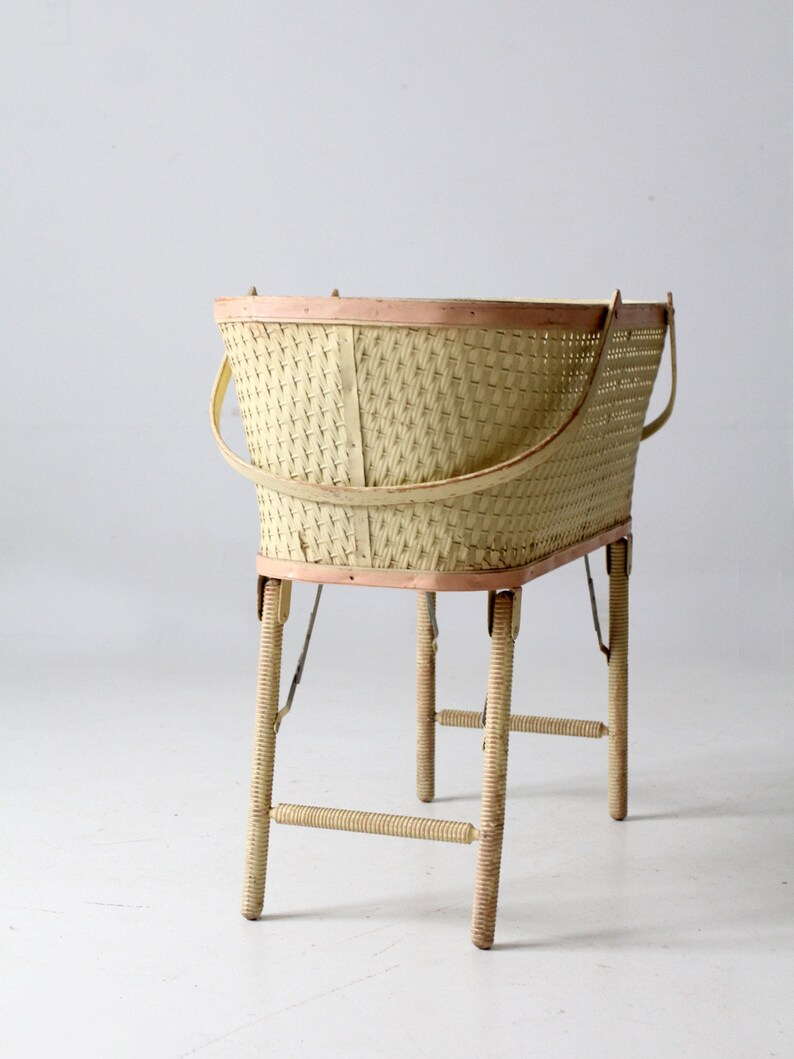May include: A vintage wicker baby bassinet with a light pink trim and wooden legs. The bassinet is on a white background.