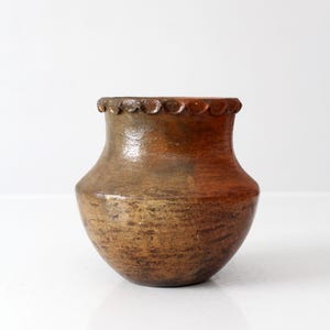 Puede incluir: Una vasija de barro redonda y bulbosa con un cuerpo ancho y un cuello estrecho. La superficie presenta un acabado moteado en tonos marrón y naranja rojizo con una apariencia texturizada. El borde está decorado con un detalle festoneado.
