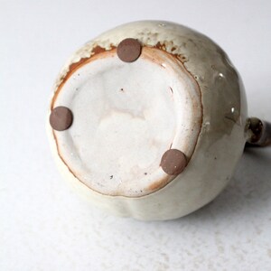 May include: The bottom of a ceramic pitcher with a light brown glaze and three brown rubber feet.
