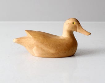 May include: A wooden duck ornament with a light brown finish. The duck has a long, flat beak, a rounded body, and small wings. The eyes are small and yellow. The ornament is on a white surface.