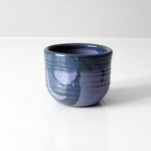 Può includere: Una piccola tazza in ceramica fatta a mano con una forma arrotondata. La tazza è dipinta con una miscela di tonalità blu e viola, con un blu più scuro all'esterno e un viola più chiaro all'interno. La tazza ha una superficie strutturata e scanalata.