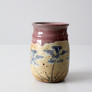 May include: A handmade ceramic vase with a cylindrical shape. The top is a deep reddish-purple, transitioning to a beige body with blue floral designs. The vase has a rustic, artisanal aesthetic.