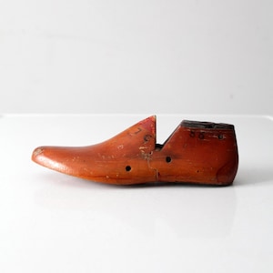 Puede incluir: Una horma de zapato de madera vintage, que muestra una forma de zapato clásica. La horma de madera es de color marrón rojizo cálido, con desgaste visible, incluyendo pequeños agujeros y una grieta en el talón. Este artículo antiguo es un testimonio de la zapatería tradicional.