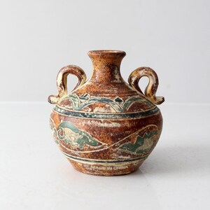 Op de afbeelding: Een kleine, bruine keramische vaas met twee gebogen handvatten. De vaas heeft een getextureerd oppervlak en is versierd met groene, blauwe en crèmekleurige bloemmotieven. De vaas heeft een ronde body en een smalle hals.