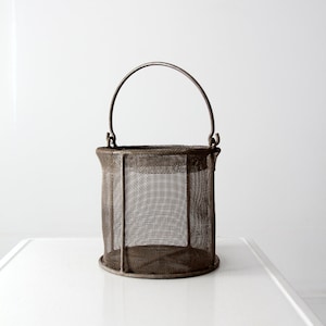 May include: Vintage metal mesh basket with a handle. The basket is cylindrical with a rounded base and a curved handle. The mesh is a dark, aged metal, and the handle is attached to the top rim. The basket is likely used for storage or display.