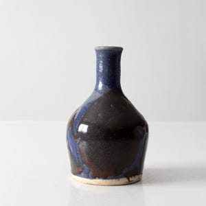 Può includere: Un piccolo vaso in ceramica con collo stretto e corpo arrotondato. La superficie presenta uno smalto lucido con un colore blu intenso sulla parte superiore e sul collo, che sfuma in una finitura marrone scuro maculata sul corpo, con una base beige non smaltata.