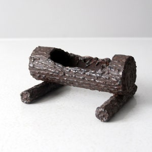 May include: A rustic, brown, log-shaped planter with a hollowed-out center. The planter is supported by two smaller log pieces, creating a unique, naturalistic design. The textured surface gives the appearance of tree bark.