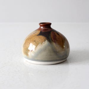 May include: A small, round ceramic vase with a narrow neck. The vase has a blend of brown, tan, and grey colours, with a glossy finish. The base is white. This decorative vase is suitable for displaying small flowers or as a standalone art piece.