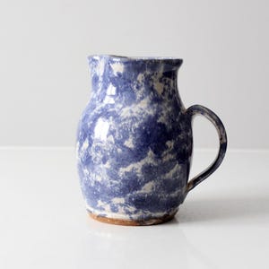 Puede incluir: Jarra de cerámica azul y blanca con asa. La jarra tiene un diseño moteado y un borde ligeramente ensanchado. La base de la jarra es de color marrón claro.