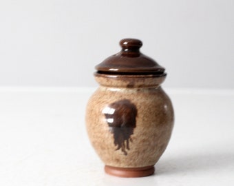 May include: A small, brown and tan ceramic jar with a lid. The jar has a brown glaze with a speckled pattern and a brown stain on the front. The lid is a simple, rounded shape.