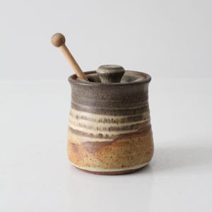 Puede incluir: Un tarro de miel de cerámica rayado en marrón y blanco con un cucharón de miel de madera. El tarro tiene una tapa con un pomo en la parte superior.