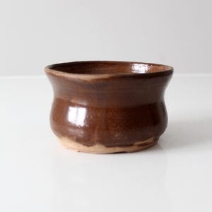 Peut inclure: Un petit bol en céramique marron avec un corps arrondi et un bord légèrement évasé. Le bol a une finition brillante et une base plus claire, non émaillée. L'intérieur est brun foncé et l'extérieur est d'une riche couleur marron chaud. Ce bol artisanal est adapté au service ou à l'exposition.