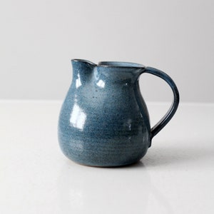 Puede incluir: Una jarra de cerámica azul con cuerpo redondeado y asa. La jarra tiene un esmalte moteado y una superficie ligeramente texturizada.