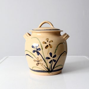 Op de afbeelding: Een beige keramische pot met een deksel, met een handgeschilderd bloemenmotief in tinten blauw, groen en bruin. De pot heeft twee handvatten aan de zijkanten en een knop op het deksel.