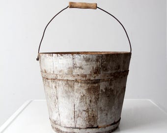 Puede incluir: Un cubo de madera antiguo y desgastado, con aros de metal y un asa de madera. El cubo tiene un aspecto rústico y envejecido, con la veta de la madera visible y un asa de metal curvada para transportarlo. Probablemente se utiliza para almacenamiento o decoración.