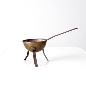 May include: A small, antique copper pot with a long handle and three legs. The pot is made of shiny, aged copper and has a rustic, vintage look.