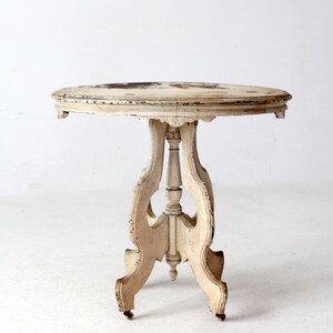 May include: A distressed, antique, round side table. The table has a cream-colored finish with visible wear and tear. It features a pedestal base with ornate, curved legs. The tabletop has a dark, mottled pattern. The table is approximately 24 inches in diameter and 28 inches tall.