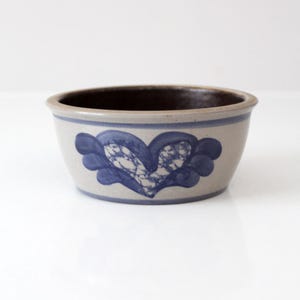 May include: A round ceramic bowl with a light beige exterior and a dark brown interior. The bowl features a blue heart-shaped design on the exterior. The rim is also blue. The bowl is likely vintage.
