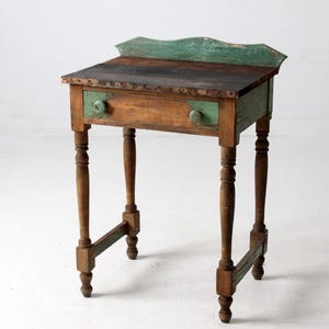 May include: Antique wooden side table with a distressed finish. The table features a green-painted top edge and drawer, with a dark brown tabletop. The drawer has two round knobs. The table stands on four turned legs and measures approximately 30 inches tall.