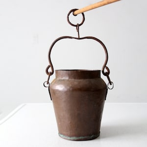 Może przedstawiać: Miedziany kubeł z metalowym uchwytem i drewnianym prętem przymocowanym do góry. Kubeł stoi na białym tle.