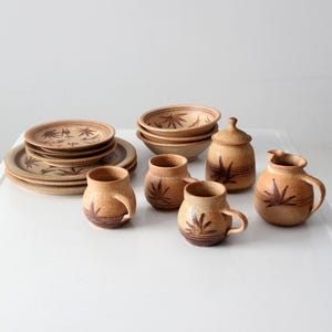 May include: A set of tan speckled ceramic dinnerware featuring dark brown painted leaf motifs. The collection includes stacked plates, bowls, three rounded mugs with handles, a lidded sugar bowl, and a small pitcher with a handle and spout.