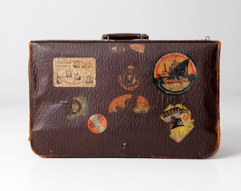 May include: Vintage brown suitcase with several travel stickers. The stickers include the text "Statler Hotels", "French Line", and "Excelsior". The suitcase has a handle and is rectangular in shape.
