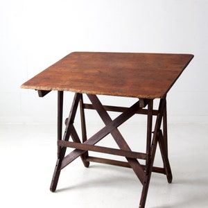 May include: A vintage wooden drafting table with a brown top and a sturdy, X-shaped base. The table is made of solid wood and has a rustic, antique look.