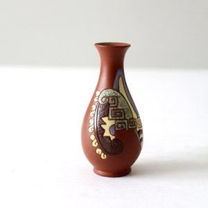 May include: A small, brown ceramic vase with a narrow neck and a rounded body. The vase is decorated with a colorful, abstract design featuring green, yellow, and brown geometric shapes and swirls. The vase is approximately 6 inches tall.