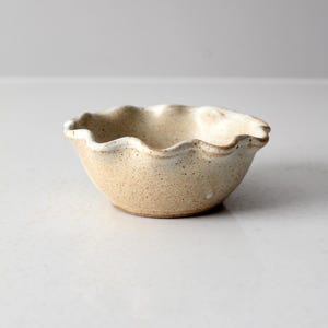 Puede incluir: Un pequeño cuenco de cerámica blanca con un borde festoneado. El cuenco tiene un esmalte beige moteado.