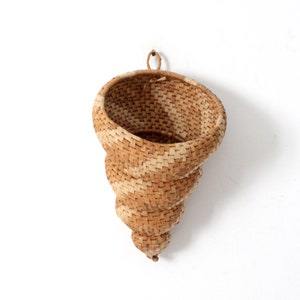 Puede incluir: Una cesta colgante tejida con materiales naturales, en tonos beige claro y crema. La cesta tiene forma de concha y tiene un asa para colgar en la parte superior.