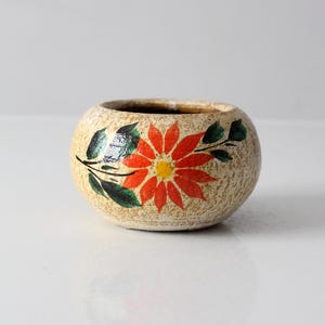 May include: A small, round, beige ceramic bowl with a textured surface. The bowl is decorated with a vibrant red and yellow flower design and green leaves. The interior of the bowl is dark brown.