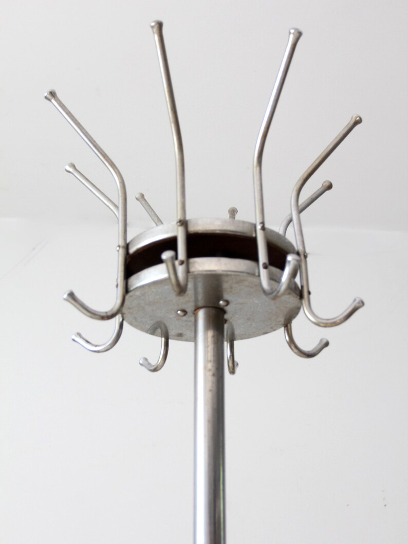 May include: A silver metal coat rack with a round base and multiple hooks. The rack is standing on a white surface.