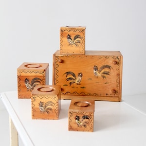 Pode incluir: Conjunto de cinco caixas de armazenamento de madeira com desenhos de galos. As caixas são pintadas em tons de marrom e apresentam ilustrações de galos pretos. A caixa maior tem uma tampa com dobradiças e as caixas menores têm tampas que se retiram.