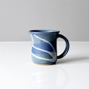 Puede incluir: Una taza de cerámica azul con diseños abstractos blancos. La taza tiene un cuerpo redondeado y un asa.