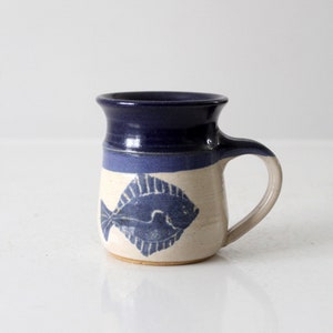 Könnte beinhalten: Eine handgefertigte Keramiktasse mit blauer und weißer Glasur. Die Tasse hat ein blaues Fischdesign auf der Vorderseite.