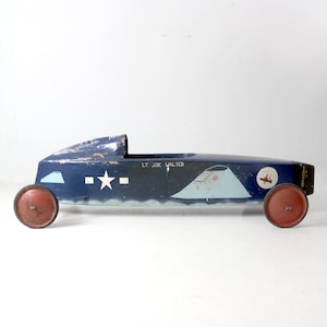 May include: A blue wooden toy race car with red wheels. The car has a white star and the text "LT. JOE WALTER" painted on the side.