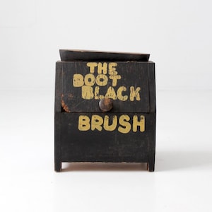 May include: A black wooden box with a hinged lid and a small round knob. The front features distressed yellow lettering that reads THE BOOT BLACK BRUSH. The box has a rectangular shape with four short legs and shows signs of wear on the paint.