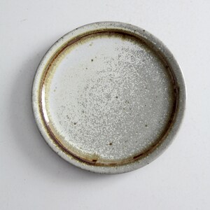 May include: A small, round, white ceramic plate with a brown rim. The plate has a speckled, textured surface.