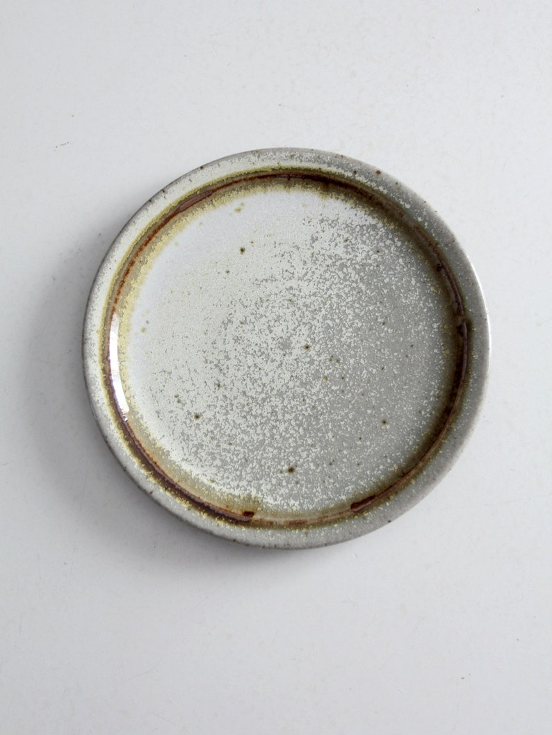 May include: A small, round, white ceramic plate with a brown rim. The plate has a speckled, textured surface.