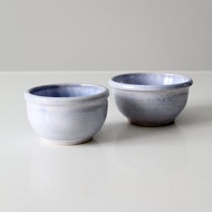 Puede incluir: Dos cuencos pequeños de cerámica azul claro con un esmalte blanco. Los cuencos son redondos y tienen un ligero borde alrededor del borde.