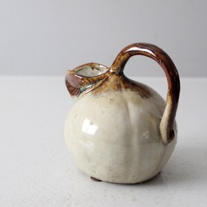 May include: A small, white ceramic pitcher with a brown handle and glaze. The pitcher has a rounded body and a small spout.