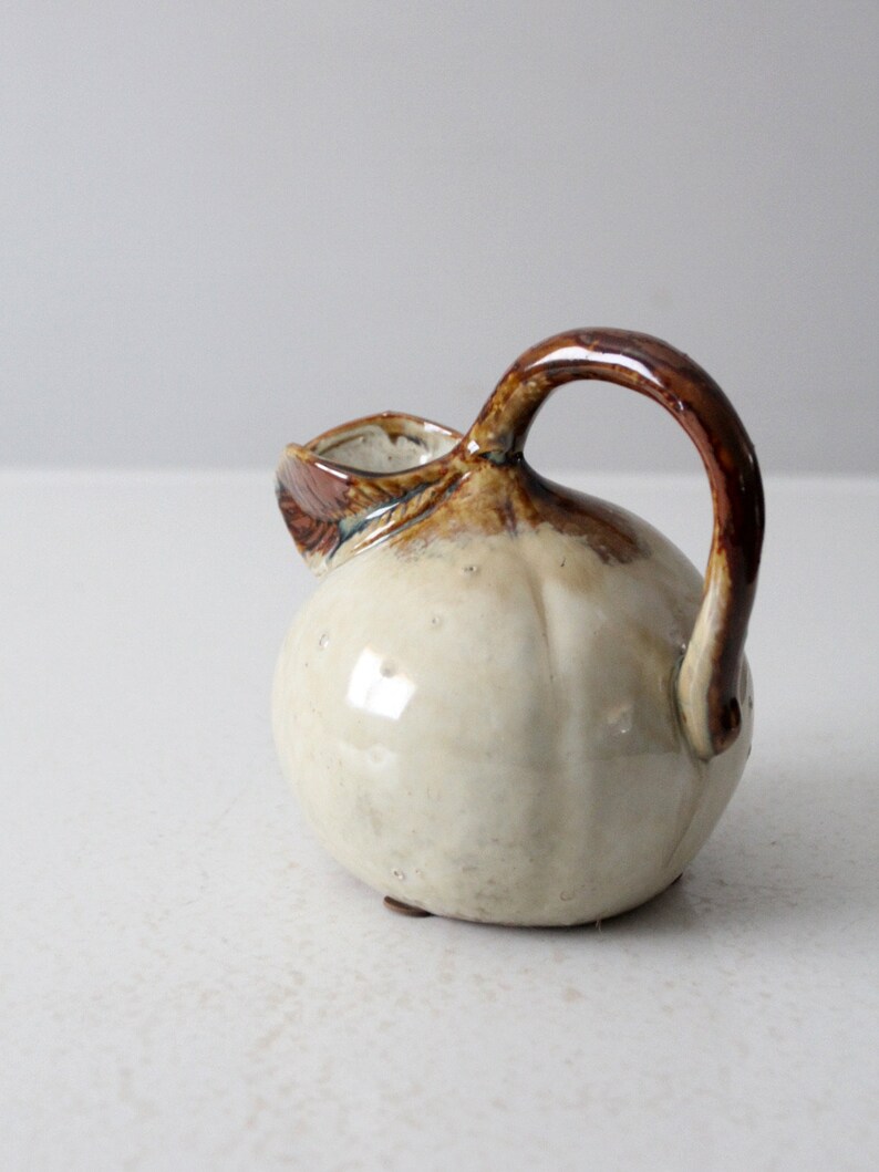 May include: A small, white ceramic pitcher with a brown handle and glaze. The pitcher has a rounded body and a small spout.