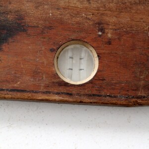 May include: A vintage wooden level with a glass bubble level. The level is brown and has a worn surface. The bubble level is clear and has a white rim.