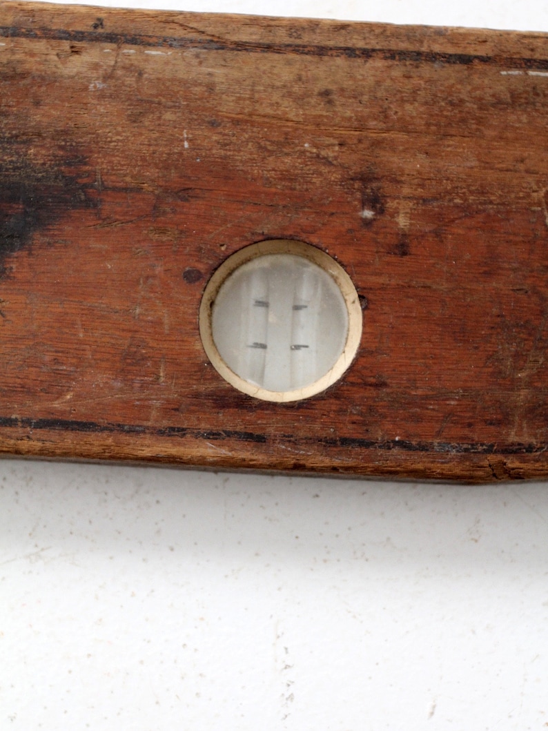 May include: A vintage wooden level with a glass bubble level. The level is brown and has a worn surface. The bubble level is clear and has a white rim.