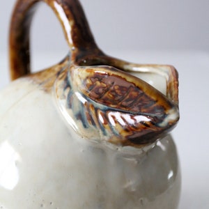 May include: A white ceramic pitcher with a brown handle and a brown leaf design. The pitcher has a glossy finish and a slightly textured surface.